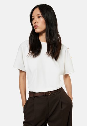 Vrouw met lang donker haar die een wit kortärmelig topje en donkerbruine broek met riem draagt, staand met haar handen in de zakken.