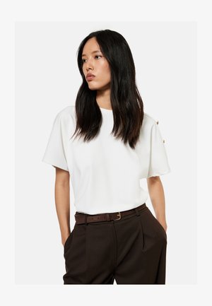 Vrouw met lang donker haar die een wit kortärmelig topje en donkerbruine broek met riem draagt, staand met haar handen in de zakken.