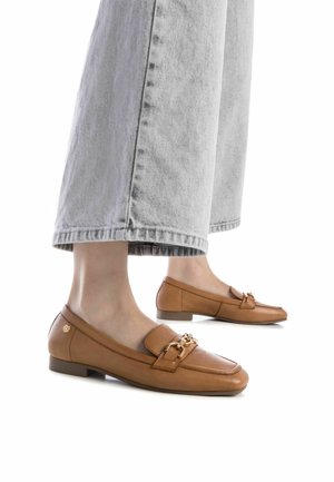 Mocasines de piel marrón con punta cuadrada, acento de cadena dorada y tacón bajo, combinados con pantalones de denim gris holgados.