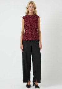 Top senza maniche color marrone granata con un design floreale testurizzato, abbinato a pantaloni neri a gamba larga e scarpe con tacco nere.