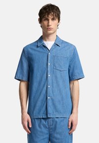 Camicia di jeans a maniche corte di colore azzurro chiaro. Presenta una chiusura con bottoni frontale, colletto e una tasca sul petto. Texture morbida con una vestibilità rilassata.