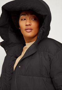 Schwarze Pufferjacke mit großer, gefütterter Kapuze aus strukturiertem Fleece, ausgestattet mit Druckknöpfen und glatter Oberfläche. Markenlabel sichtbar auf der Brust.