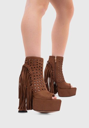 Stivaletti marroni in suede con plateau, tacco alto, dettagli traforati, cerniera laterale, punta aperta e lunghe frange sui lati esterni, indossati a gambe nude.