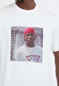 Biała bawełniana koszulka z dużym nadrukiem w postaci zdjęcia, na którym znajduje się czerwony trójkątny logo oraz portret osoby noszącej czerwoną czapkę i naszyjnik.