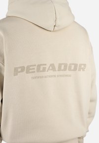 Beige hettegenser med teksturert stoff, med en stor trykt logo "PEGADOR" i en slitt skrifttype og "CERTIFIED AUTHENTIC STREETWEAR" under.