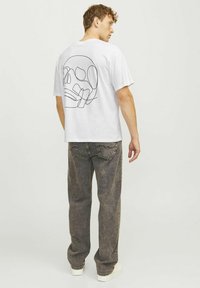 Hvid bomulds t-shirt med en sort stregtegning af et kranium på ryggen, parret med grå, let baggy denimjeans og hvide sneakers.