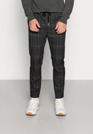 Man wearing dark gray plaid trousers with a drawstring waist, white sneakers, and a dark long-sleeve top, standing on a gray floor.
