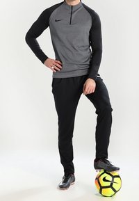 Camiseta de manga larga gris con cremallera de cuarto, con mangas negras, combinada con pantalones deportivos negros. Un balón de fútbol con acentos en amarillo neón y negro está bajo los pies.