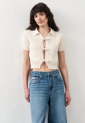 Femeie cu păr creț și închis la culoare, purtând un cardigan tricotat crem, cu mâneci scurte și trei fundițe în față, și blugi albaștri cu talie înaltă.