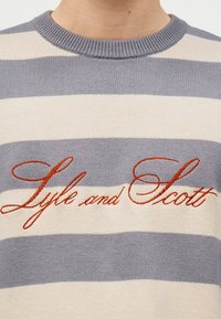 Κοντινή λήψη γκρι και λευκού ριγωτού πλεκτού πουλόβερ με κεντημένο το «Lyle and Scott» σε πορτοκαλί καλλιγραφικά πάνω στο στήθος.