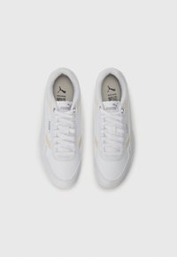 Baskets de sport blanches avec des lacets plats, tissu texturé, une bande d'accent dorée légère et une semelle en caoutchouc. Logo Puma sur la languette et sur le côté.