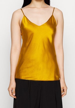 Kobieta ubrana w żółty satynowy top na cienkich ramiączkach bez rękawów w kolorze musztardowym oraz czarne spodnie, przedstawiona od ramion do bioder.