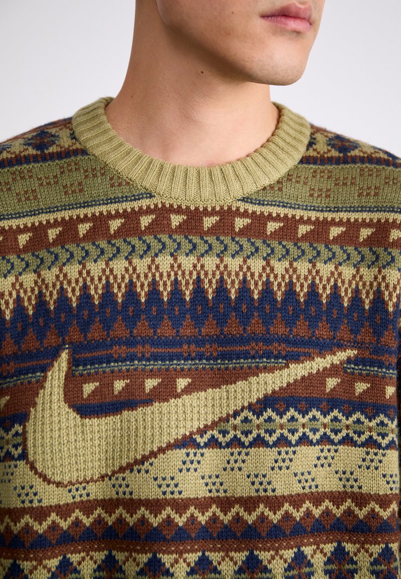 Un pull en tricot avec une base vert olive, des accents à motifs marron et bleu, un col rond et un logo Nike doré bien en vue.