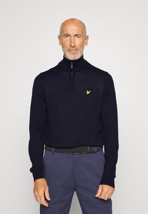 Pull en maille marine avec un col haut et une fermeture à demi-zip. Présente un petit logo jaune sur la poitrine et des poignets côtelés. Porté avec un pantalon foncé.