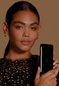 Smartphone avec une finition noire élégante tenu contre une tenue noire texturée avec des accents dorés. Le mannequin a des cheveux mi-longs et sombres ainsi que des boucles d'oreilles.