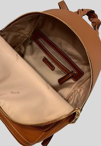 Zaino in pelle tan con chiusura a zip. L'interno presenta una fodera beige, una tasca con zip e il logo del marchio stampato su tutta la superficie. Accenti in metallo dorato.