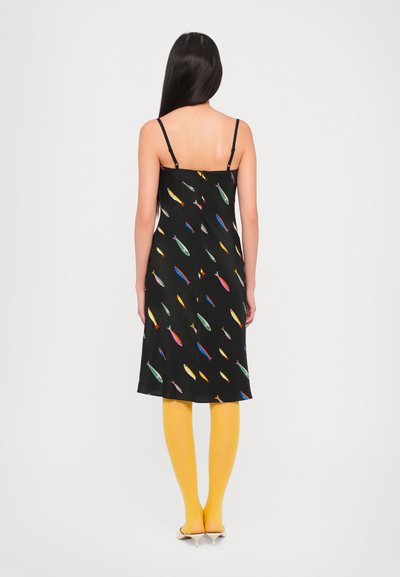 Zwarte slipdress met een multicolor vissenprint, verstelbare bandjes, middellange snit, gecombineerd met gele panty’s en puntige hakken.