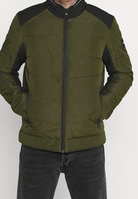 Grøn puffjakke med sorte skulderdetaljer, lynlås foran, opretstående krave og to forlommer; quiltet struktur.