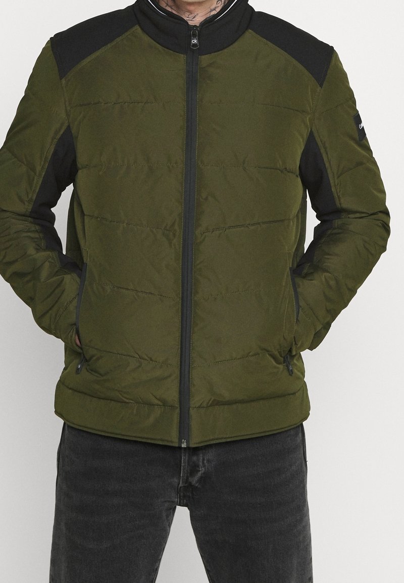 Grøn puffjakke med sorte skulderdetaljer, lynlås foran, opretstående krave og to forlommer; quiltet struktur.