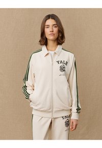 Cream zip-up jacket with green stripes, "Yale" graphic and bulldog logo on the chest, featuring a relaxed fit and soft texture.