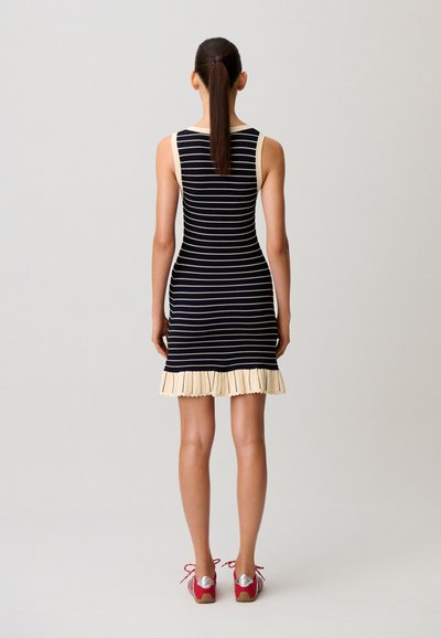 Frau mit langem braunem Pferdeschwanz, die ein ärmelloses schwarz-weiß gestreiftes Kleid mit cremefarbenem Rüschensaum und roten Turnschuhen trägt, steht mit dem Rücken zum Betrachter vor neutralem Hintergrund.