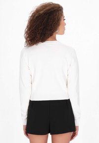 Fehér kötött pulóver hosszú ujjakkal és bordás aljjal, fekete sorttal párosítva. A hátulról készült nézet egy egyszerű, klasszikus dizájnt és textúrát mutat.