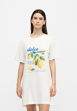 Frau trägt ein weißes oversized T-Shirt mit einem Zitronenzweig und italienischem Textdruck, steht vor einem schlichten hellgrauen Hintergrund.