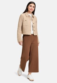Giacca beige corta con trama tessuta, chiusure a bottone e orlo grezzo. Abbinata a pantaloni larghi marroni e sneakers bianche.