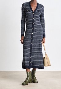 Námořnické pruhované pletené šaty s dlouhými rukávy, V-výstřihem, zlatými knoflíky aá přiléhavým střihem. Doplňky zahrnují béžovou kabelku a zelené kotníkové boty.