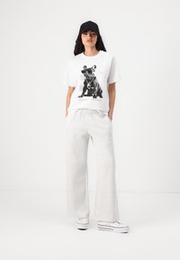 White graphic T-shirt featuring a black dog illustration, paired with light grey wide-leg sweatpants and white sneakers. Black cap worn.