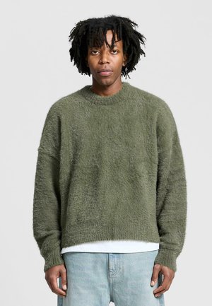 Olivgrüner, flauschiger Strickpullover mit Rundhalsausschnitt, überschnittenen Schultern und gerippten Bündchen. Über einem weißen Hemd getragen und mit hellen Jeans kombiniert.
