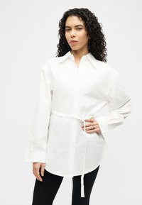 Γυναίκα με κατσαρά μαλλιά που φοράει λευκό πουκάμισο με ζώνη και μακρύ μανίκι και μαύρο παντελόνι, πόζαρε με το ένα χέρι στη μέση της μπροστά σε λευκό φόντο.