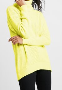 Nainen, jolla on yllään kirkkaan keltainen paksu neulepusero, jossa on pitkät hihat, ja mustat housut, poseeraa yksi käsi kasvojensa lähellä.