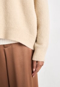 Beige ulltröja med ribbade mudd och nederkant, som har en avslappnad passform. Tillsammans med släta, bruna tygbyxor. Mjuk textur synlig.