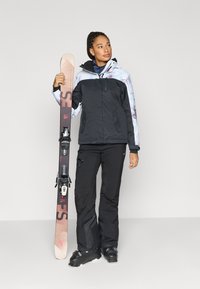 Slēpošanas jaka ar rakstainu gaiši zilu un melnu dizainu, kopā ar melnām slēpošanas biksēm un melniem slēpošanas zābakiem, turot slēpes ar stiprinājumiem.