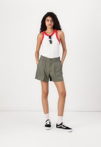 Vit linne med röd kant, khakishorts och svarta sneakers. Modellen har solglasögon, har lockigt hår, står med händerna i shortsfickorna.
