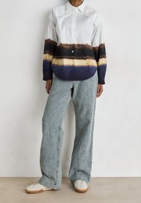 Persona con una camisa de botones de colores múltiples con rayas marrones, negras, amarillas y azules, jeans de pierna ancha azul claro y zapatillas blancas.