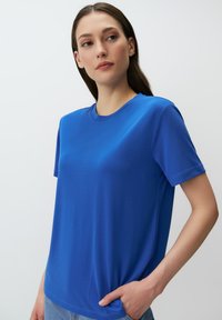 Niebieska bawełniana koszulka z krótkimi rękawami i okrągłym dekoltem. Gładka tekstura i luźny krój, z ręką modela w kieszeni.