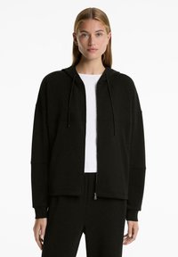 Žena s rovnými blond vlasy oblečená v černé mikině na zip a odpovídajících kalhotách přes bílé tričko, stojící na bílém pozadí.