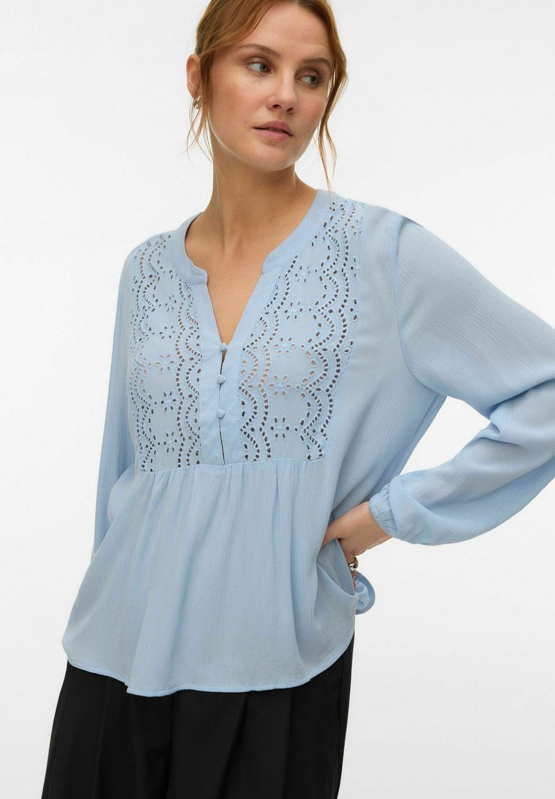 Femme portant un chemisier bleu clair à manches longues avec broderie ajourée festonnée et boutons recouverts de tissu, rentrée dans un pantalon noir.