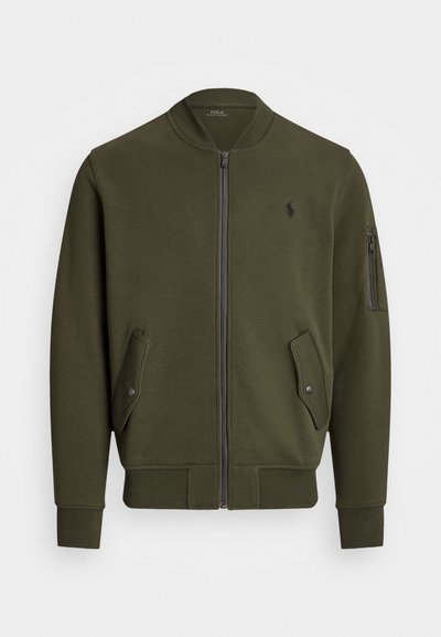 JAqueta bombere verde-azeitona em tecido suave com fecho de zip, dois bolsos laterais e um bolso com zip na manga. Apresenta um logótipo no peito.