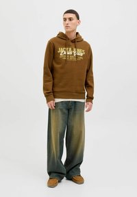 Brun hættejakke med grafisk tekst på forsiden, parret med vide, falmede blå jeans og brune slip-on sko. Glat stof, afslappet pasform.