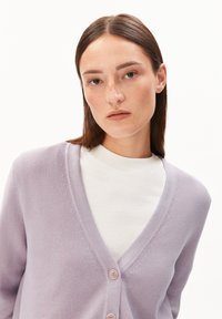 Lavender cardigan van zacht breiwerk, met een diepe V-halslijn en twee knopen aan de voorkant. Draagt over een wit shirt.
