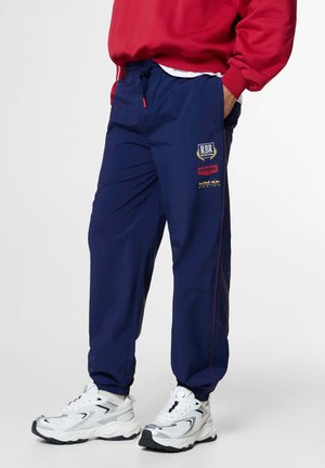 Person, die dunkelblaue Red Bull Racing-Hosen und weiße Sneakers trägt, mit den Händen in den Taschen und einem teilweise sichtbaren roten Sweatshirt.