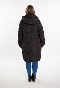 Černá péřová bunda s kapucí, se šitým vzorem srdcí. Modelka má na sobě modré džíny a černé kotníkové boty.