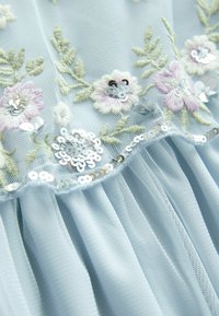 Lyseblåt gennemsigtigt stof med broderede blege lilla og hvide blomster, grønne blade og sølv palietter langs en flæsekant.