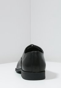 Chaussure habillée en cuir noir avec une texture lisse, un bout rond et un talon bas. Présente une couture le long de l'arrière et aucune décoration visible.