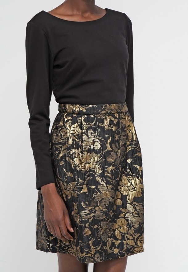 Femme portant un haut noir à manches longues associé à une jupe taille haute avec un motif floral noir et or.