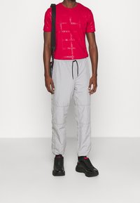 Rotes T-Shirt mit glänzendem Logo-Print, kombiniert mit leichten, hellgrauen Hosen mit Kordelzug und Seitentaschen. Schwarze Sneakers.