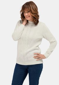 Frau mit schulterlangem braunem Haar, die einen weißen Strickpullover und blaue Jeans trägt, lächelt und hat eine Hand am Hals und die andere auf der Hüfte.
